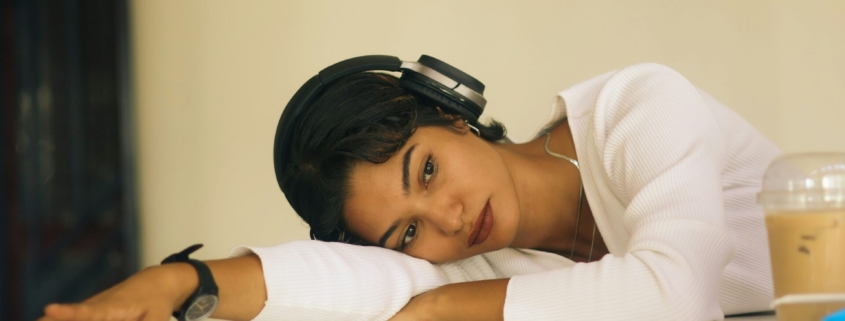 A young South Asian woman with headphones on laying her torso on a table indoors, looking distressed.