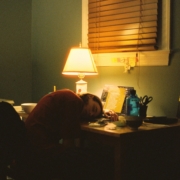 A person sleeping at a desk in a dark room in front of a lamp.
