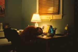 A person sleeping at a desk in a dark room in front of a lamp.