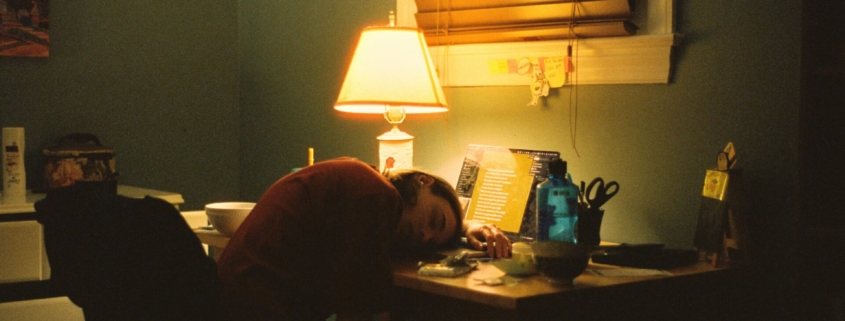 A person sleeping at a desk in a dark room in front of a lamp.