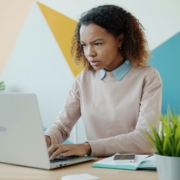 A Black woman at desk looking at a computer in front of a painted wall, looking stressed.