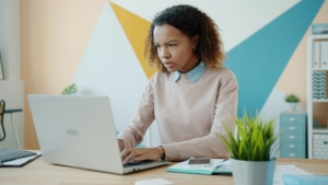 A Black woman at desk looking at a computer in front of a painted wall, looking stressed.