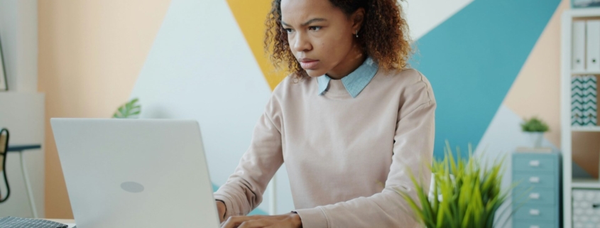 A Black woman at desk looking at a computer in front of a painted wall, looking stressed.